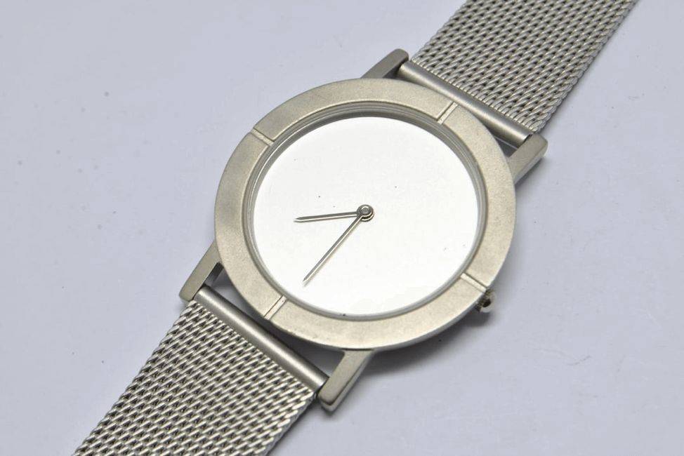 A top-down shot of a minimalist watch with a plain white face and two silver hands. The watch has a silver-tone metal casing and a fine mesh metal strap. The watch face is blank without any numbers making it hard to tell the time, it has a raised circular border and four small, indented marks at the 12, 3, 6, and 9 o'clock positions. The watch is positioned diagonally across a white surface.
